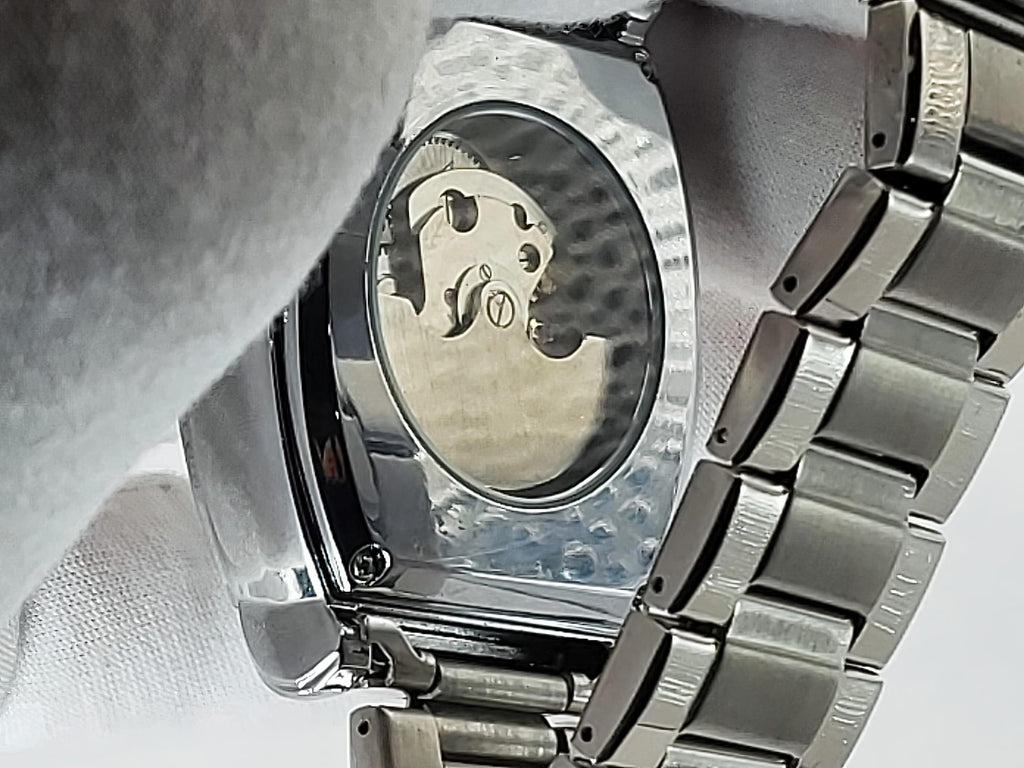 Close-up of the back of a stainless steel Aokulasic Open Heart Automatic watch with a transparent case back showing the internal movement, held by a white-gloved hand.
