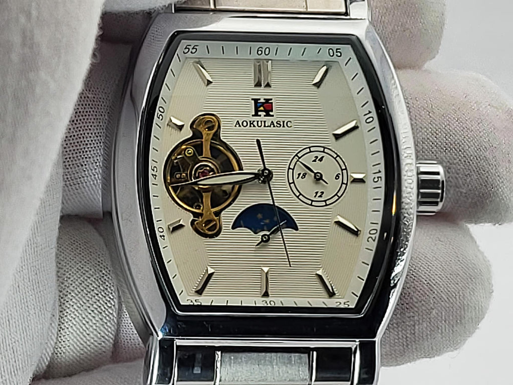 A close-up of an Aokulasic Open Heart Automatic men's watch with a silver-tone stainless steel case and a cream-colored dial, held by a hand wearing a white glove. The dial features a visible gold-tone mechanical movement, a moonphase indicator, and a small seconds sub-dial.