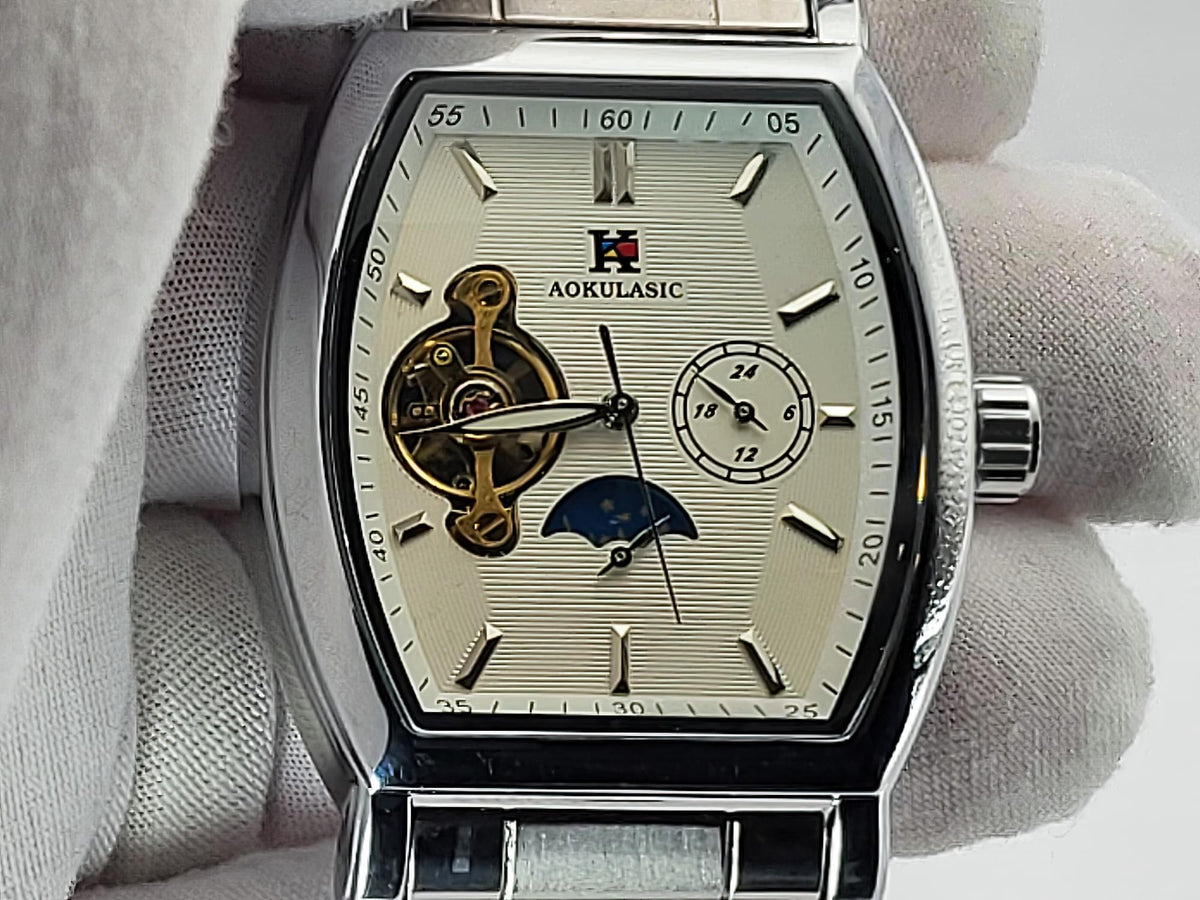 A close-up of an Aokulasic Open Heart Automatic men's watch with a silver-tone stainless steel case and a cream-colored dial, held by a hand wearing a white glove. The dial features a visible gold-tone mechanical movement, a moonphase indicator, and a small seconds sub-dial.
