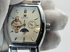 A close-up of an Aokulasic Open Heart Automatic watch with a silver-tone case and a cream-colored dial, held by a hand wearing a white glove. The dial features a skeletonized design showing the inner movement, a moon phase indicator, and a 24-hour sub-dial.