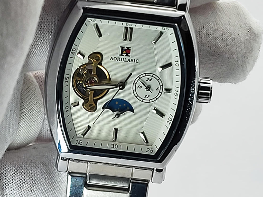 Aokulasic Open Heart Automatic men's watch with a silver-tone stainless steel case, featuring a white dial with skeletonized hour markers, a gold-toned automatic movement, and a blue moon phase indicator, held by a white-gloved hand.