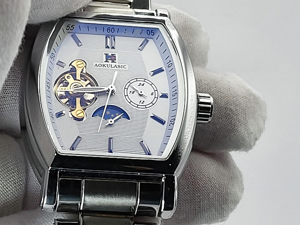 A close-up of a silver-toned Aokulasic Open Heart Automatic men's watch with a silver dial, blue hands and markers, and a visible gold-toned mechanical movement, held by a white gloved hand.