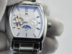 A close-up of a silver-toned Aokulasic Open Heart Automatic men's watch with a silver dial, blue hands and markers, and a visible gold-toned mechanical movement, held by a white gloved hand.