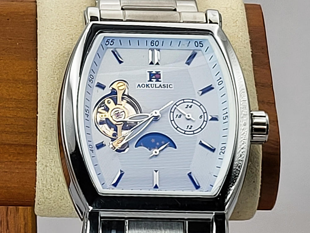 A close-up of an Aokulasic Open Heart Automatic men's watch with a silver stainless steel case, a light blue dial featuring a skeletonized movement and moonphase indicator, and blue hour markers, displayed on a beige cushion mounted on a wooden stand.