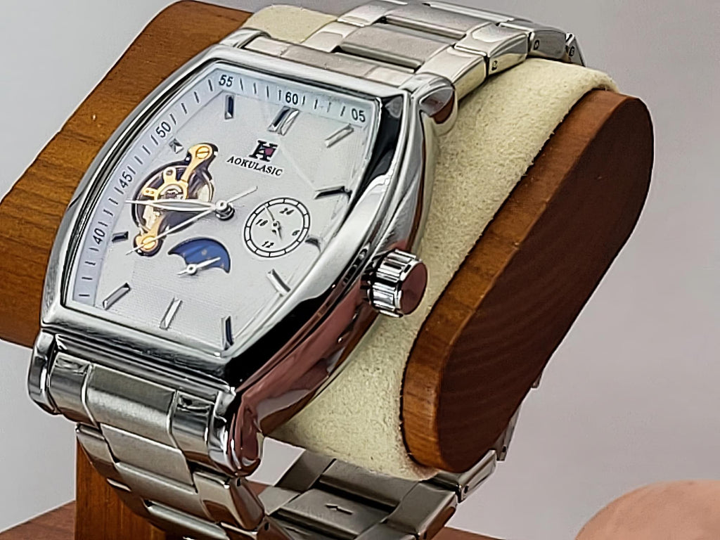 A silver-toned Aokulasic Open Heart Automatic watch with a rectangular rounded case, silver link bracelet, and a white dial featuring a moonphase indicator and small subdial, displayed on a wooden stand with a cream cushion.