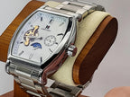 A silver-toned Aokulasic Open Heart Automatic watch with a rectangular rounded case, silver link bracelet, and a white dial featuring a moonphase indicator and small subdial, displayed on a wooden stand with a cream cushion.