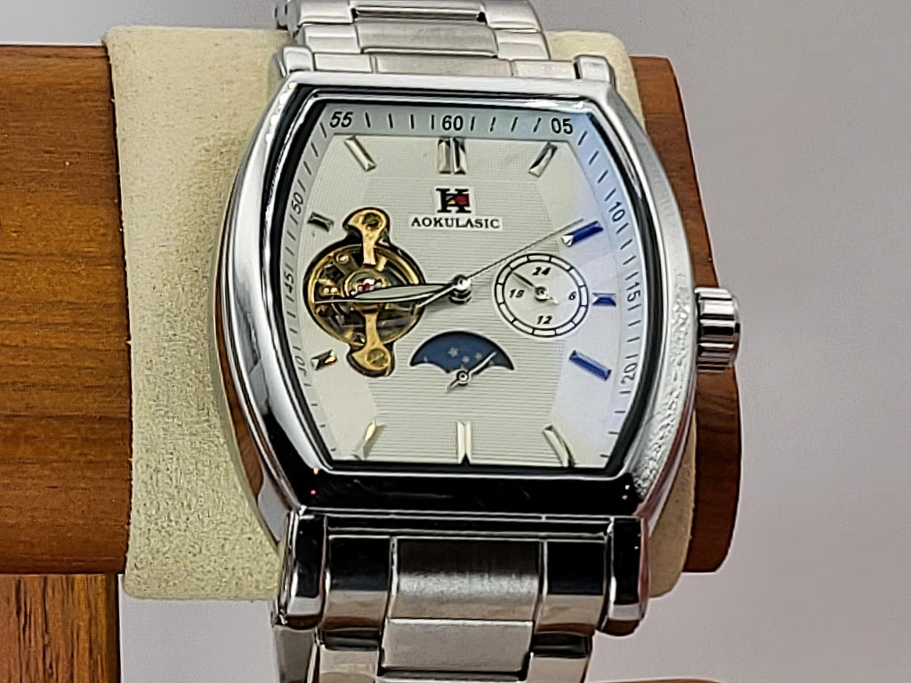 Aokulasic Open Heart Automatic men's automatic watch with a silver stainless steel case and link bracelet, featuring a white dial with skeletonized details, blue hands, and a moon phase indicator, displayed on a wooden stand.