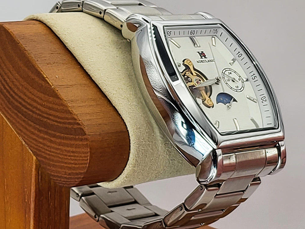 A close-up of a Aokulasic Open Heart Automatic wristwatch with a polished silver-tone metal case and link bracelet, mounted on a beige display cushion inside a wooden holder. The watch has a white dial with skeletonized details showing internal gears and a blue moonphase indicator.