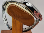 A close-up of a silver stainless steel Aokulasic Open Heart Automatic watch band with a polished chrome finish, mounted on a wooden display stand with a cream-colored foam insert.