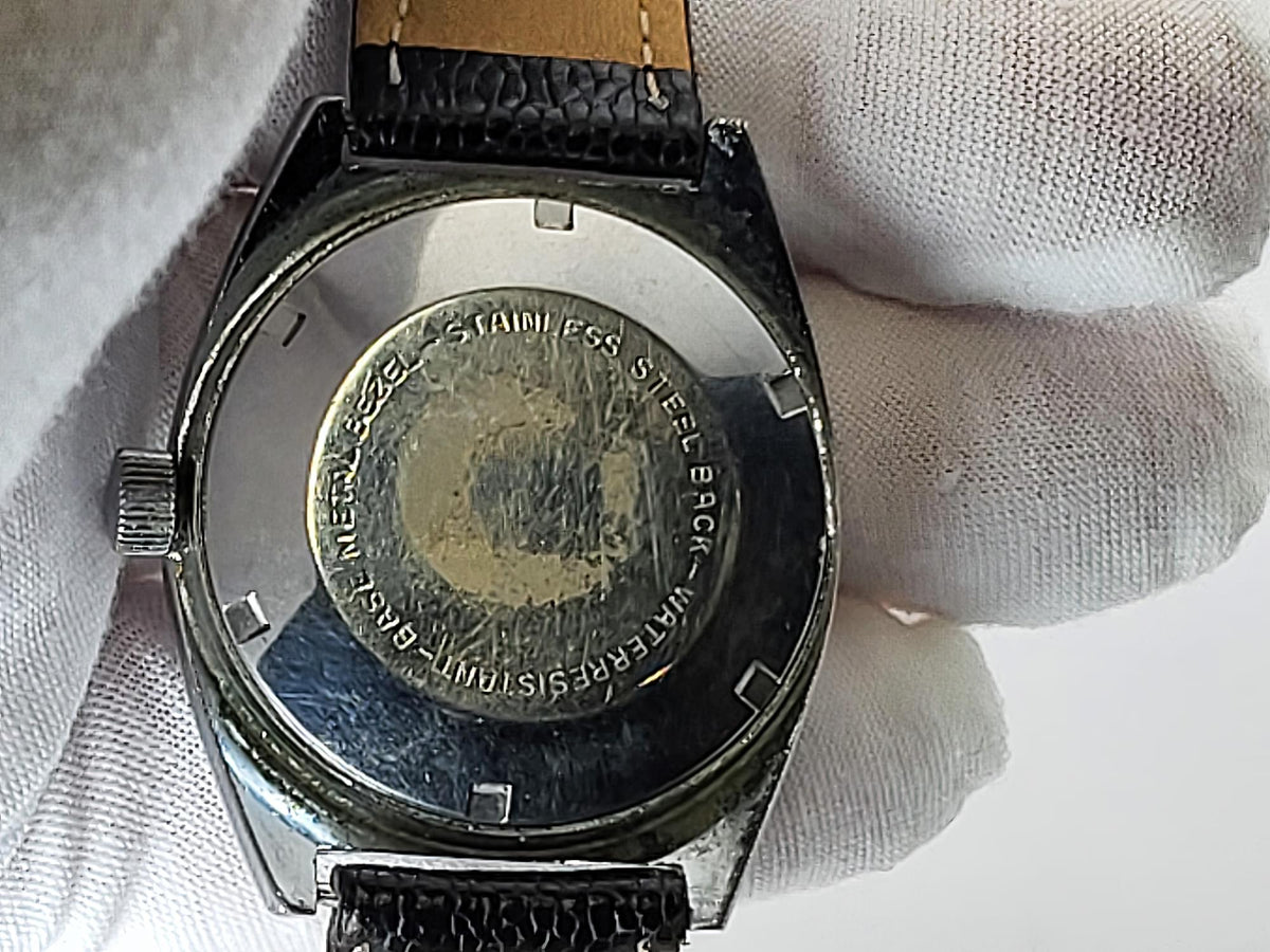 Close-up of the back of a vintage stainless steel Benrus wristwatch held by a white gloved hand. The watch has a black leather strap and a brushed metal case back with engraved text.