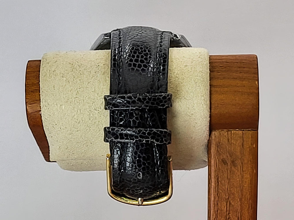 A Benrus wristwatch black embossed leather watch strap with a gold buckle, displayed on a cream-colored foam roll mounted on a wooden stand against a light gray background.