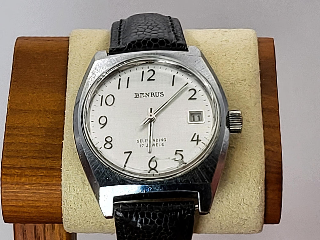 A vintage Benrus wristwatch with a silver-toned metal case, white dial, and black leather strap, displayed on a beige cushion inside a wooden stand.