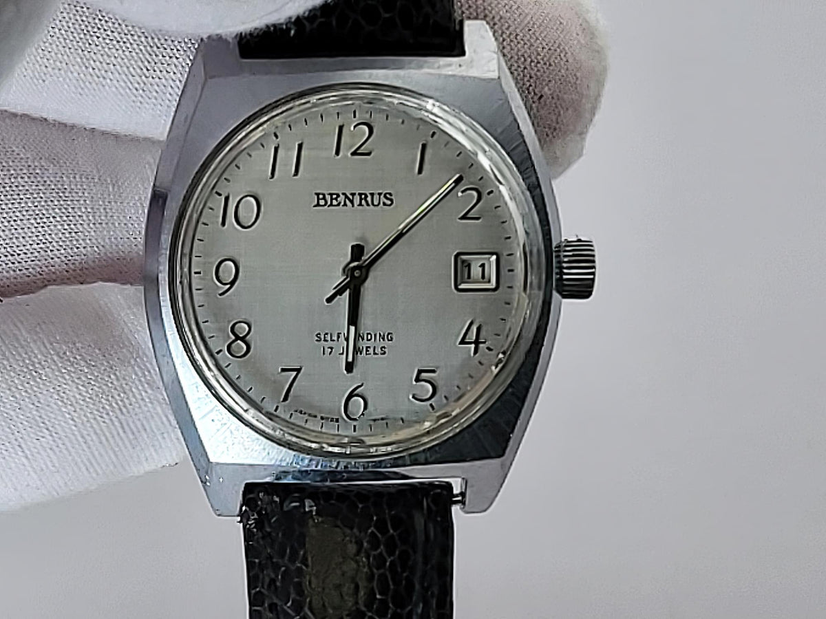 A vintage Benrus wristwatch with a silver-toned metal case, white dial, and black textured strap, held by a white-gloved hand against a light gray background.