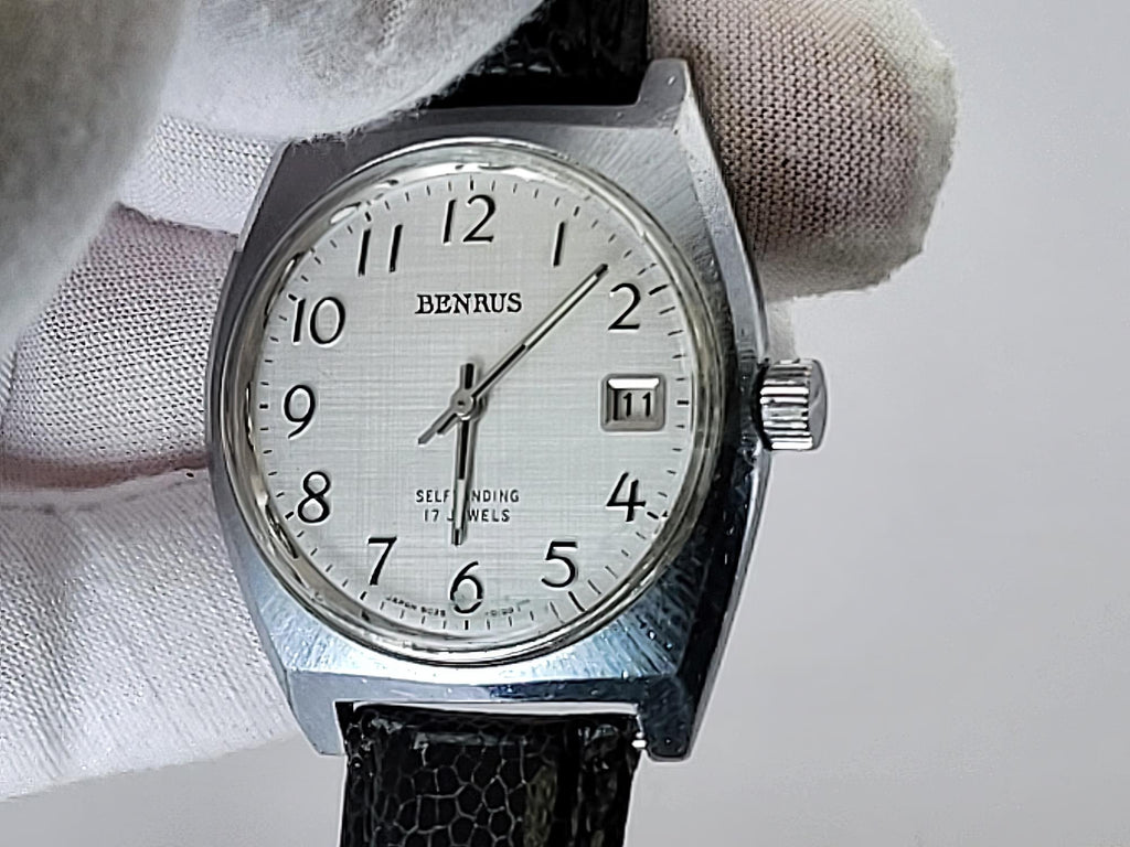 A vintage Benrus wristwatch with a silver-toned metal case, white dial, and black leather strap, held by a white gloved hand against a light gray background.