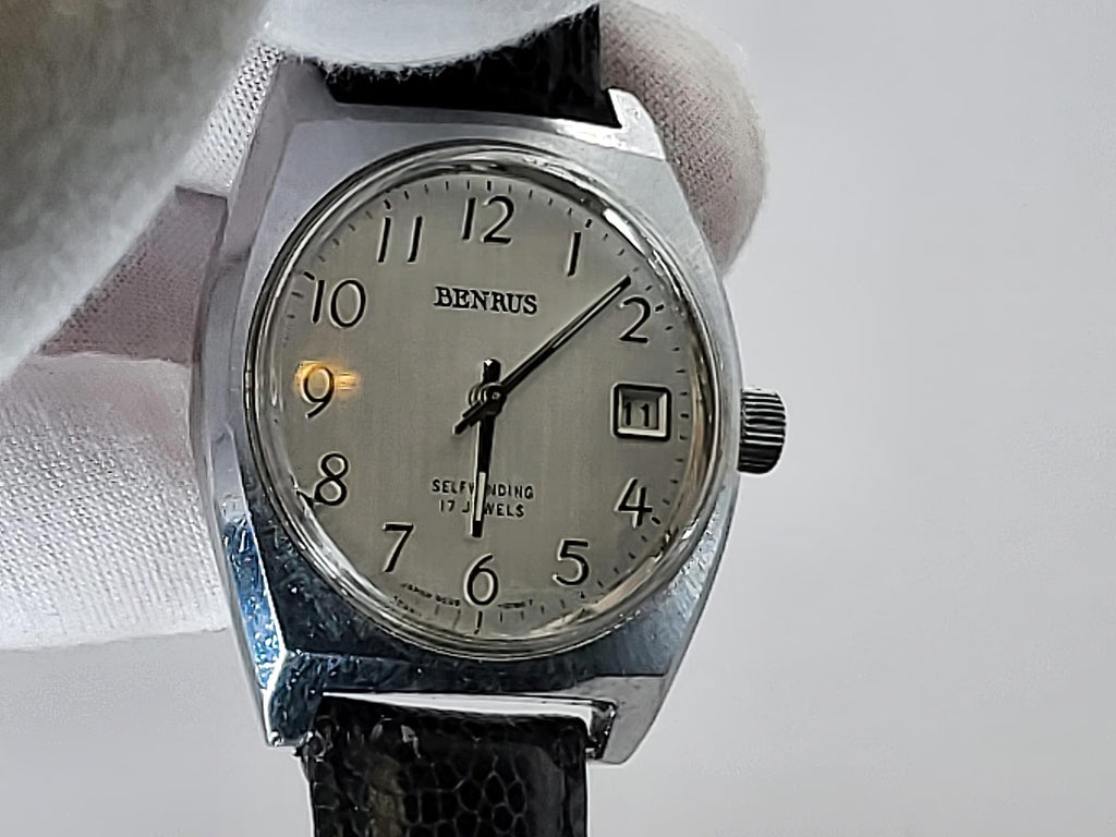 A vintage Benrus watch with a silver-toned metal case, a cream-colored dial, and a black leather strap, held by a white gloved hand against a light gray background.