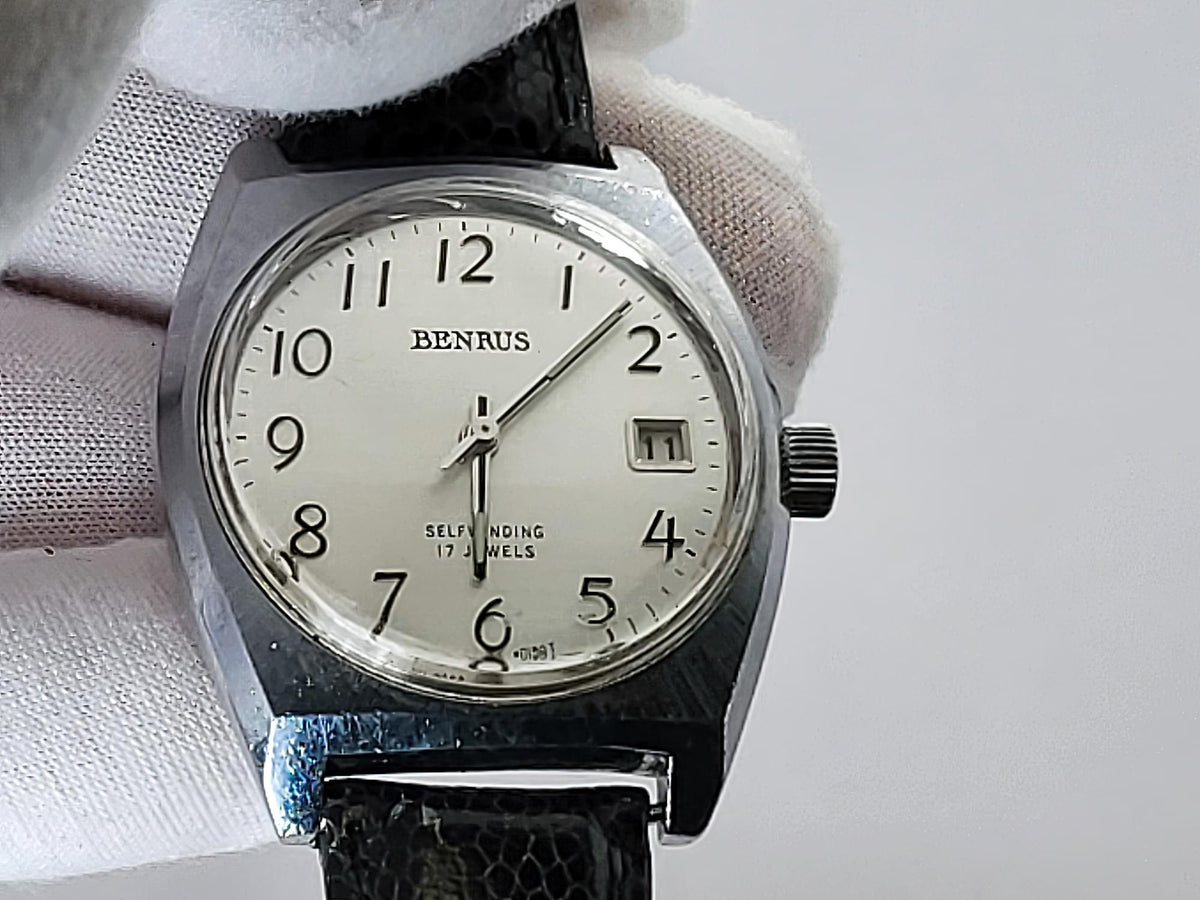 A vintage Benrus wristwatch with a silver-toned metal case, white dial, and black leather strap, held by a white gloved hand against a light gray background.