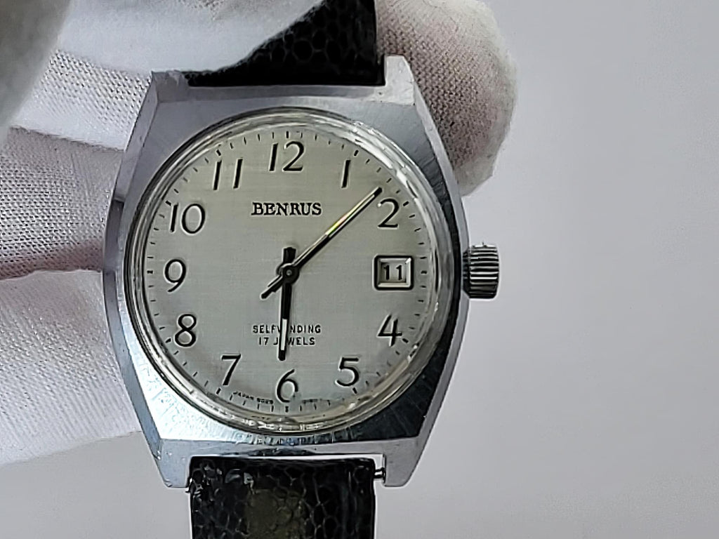 A vintage Benrus wristwatch with a silver-toned metal case, cream-colored dial, and black leather strap, held by a white-gloved hand against a light gray background.