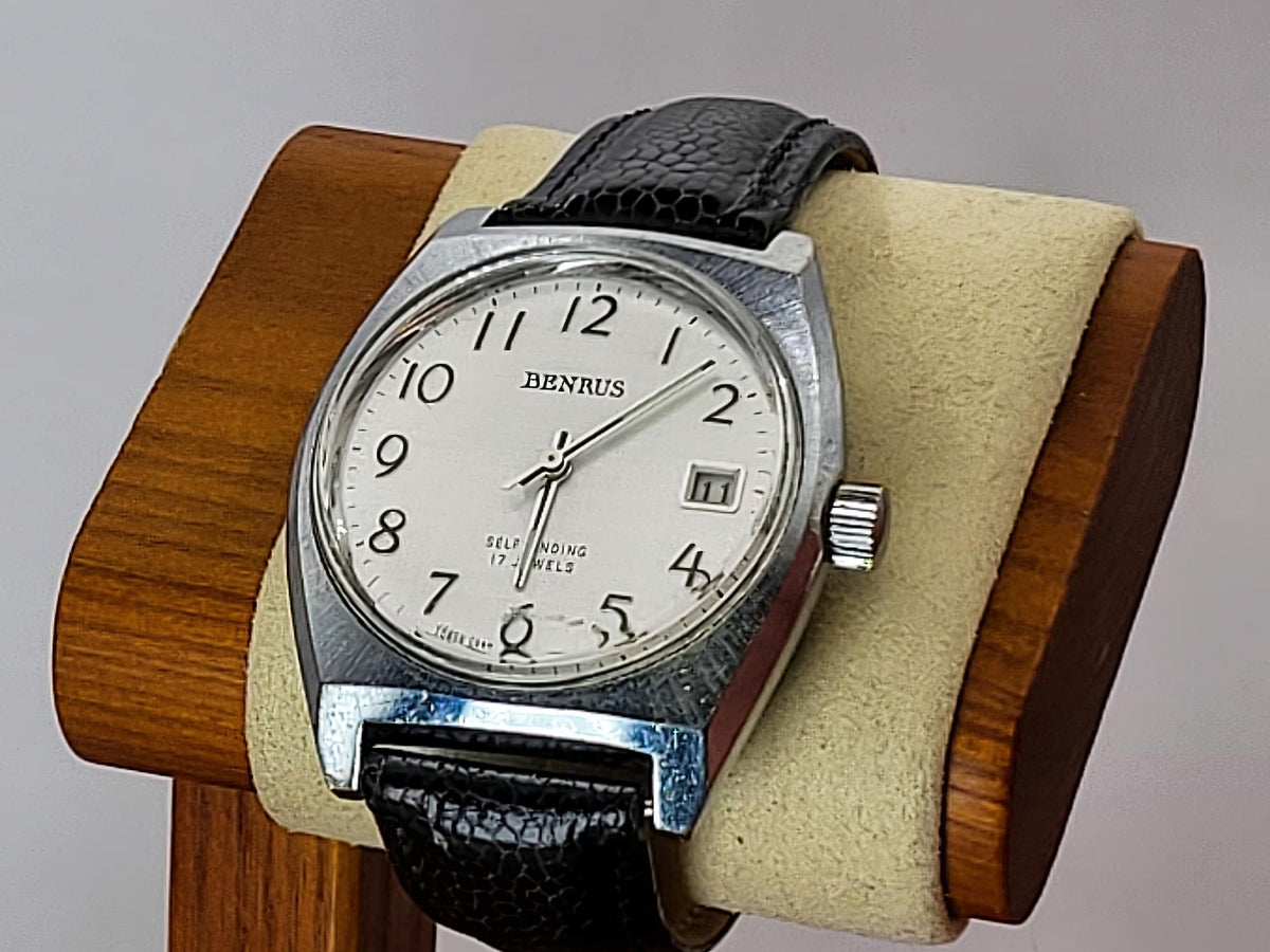 A Bernus wristwatch with a silver-toned case, white dial, and black leather strap, displayed on a beige cushion inside a wooden stand against a neutral gray background.