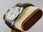 A Benrus watch with a silver-tone case, white dial, and black leather strap, displayed on a cushioned wooden stand against a light gray background.