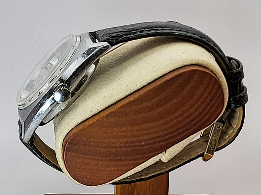 A vintage silver-tone Benrus wristwatch with a black leather strap, displayed on a wooden stand with a cream-colored cushion. The watch is photographed against a neutral light gray background.