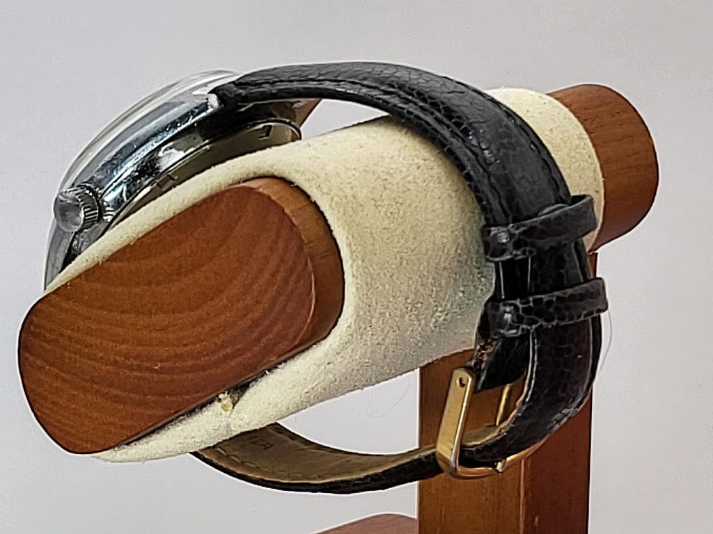 A vintage Benrus wristwatch with a black leather strap and silver-toned case, displayed on a wooden stand with a cream-colored velvet cushion.