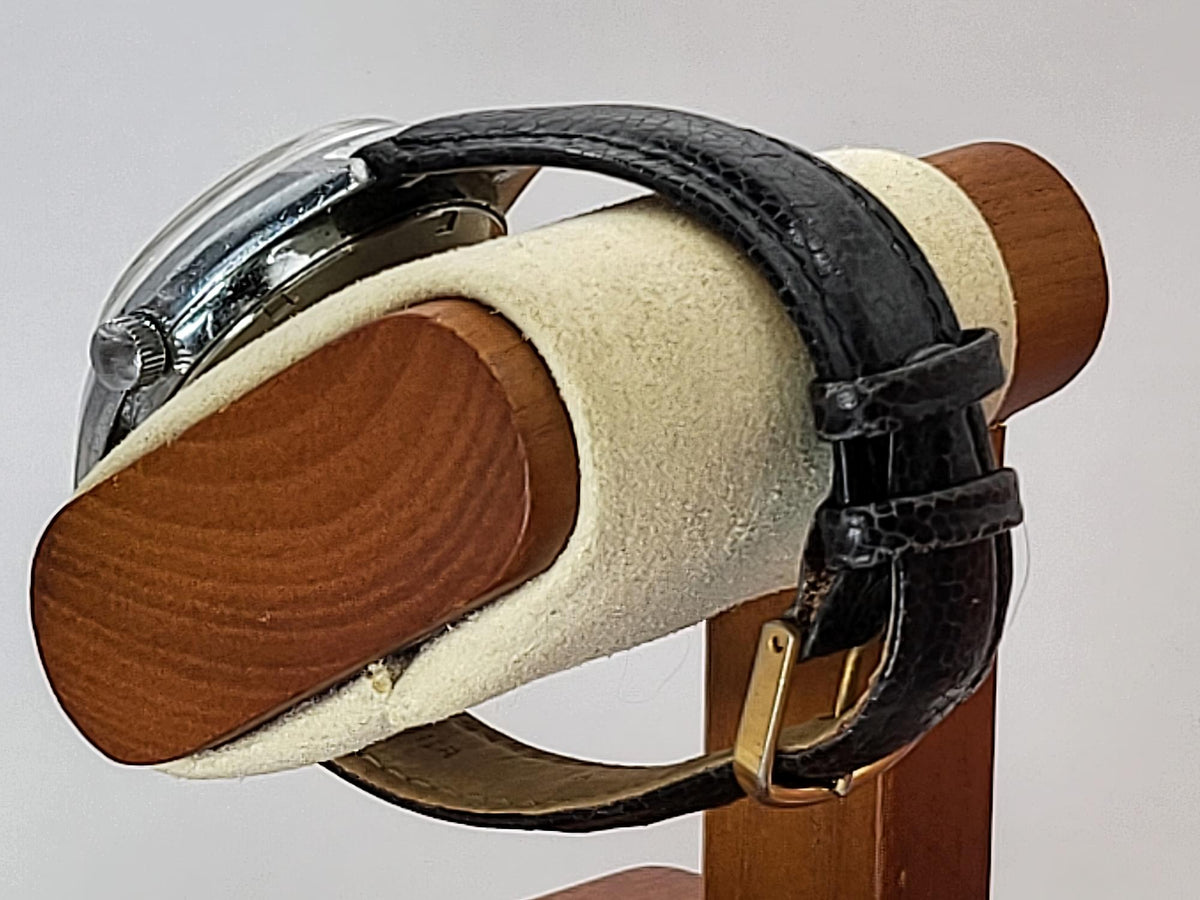 A vintage Benrus wristwatch with a black leather strap and silver-toned case, displayed on a wooden stand with a cream-colored velvet cushion.