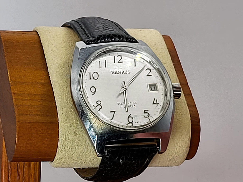 A Benrus wristwatch with a silver-toned case, white dial, and black leather strap, displayed on a beige cushion mounted on a wooden stand against a light gray background.