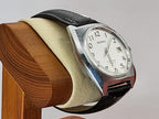 A vintage Benrus brand wristwatch with a silver-tone case, white dial, and black leather strap, displayed on a beige cushion mounted on a wooden stand against a light gray background.