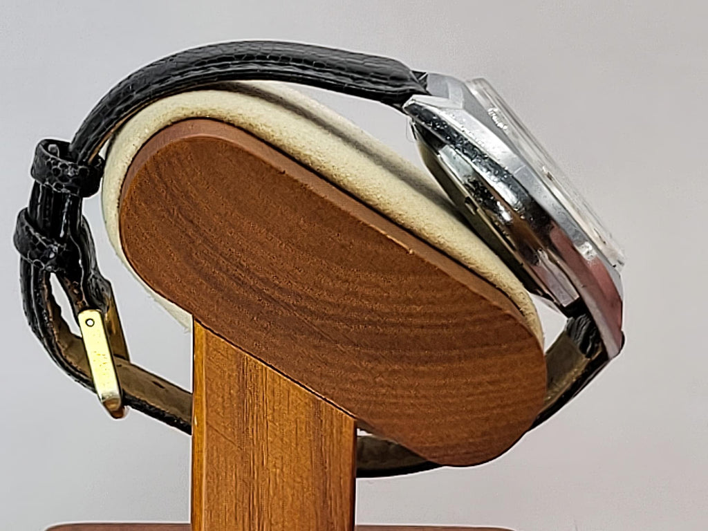 A Benrus wristwatch with a black leather strap and silver-toned case, displayed on a wooden stand against a light gray background.