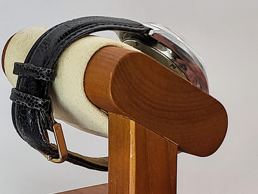 A close-up of a Benrus wristwatch with a black textured leather strap and a silver-toned round case, displayed on a cream cylindrical stand with a wooden arm against a light gray background.