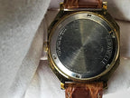 Close-up of the back of a vintage Caravelle Day-Date wristwatch with a gold-tone case, brushed silver metal back cover, and brown leather strap resting on a white textured fabric.