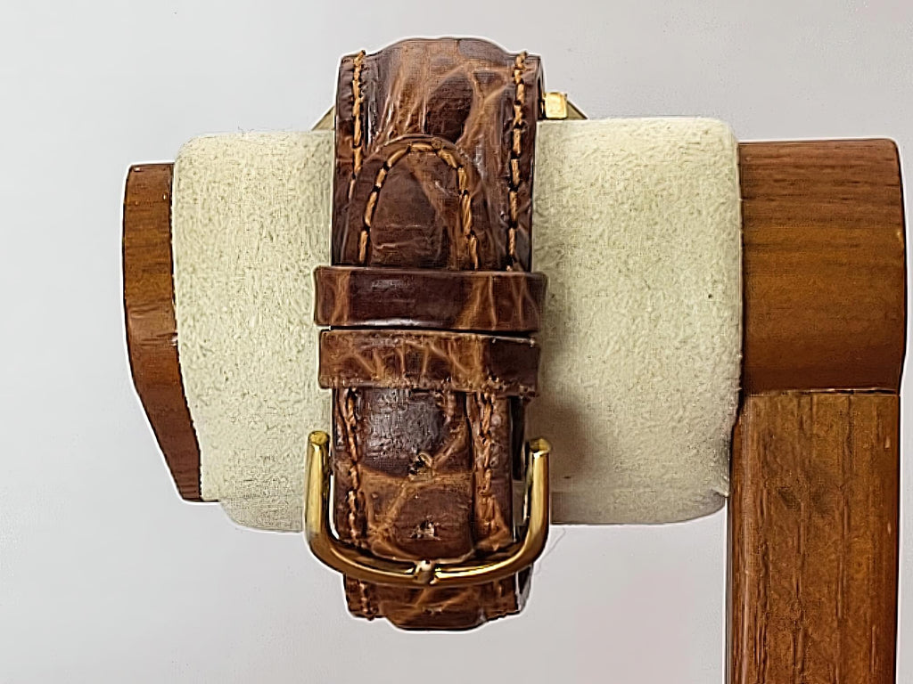 A brown crocodile-embossed leather Caravelle Day-Date watch strap with gold-tone hardware, displayed on a cream-colored velvet watch stand with a wooden base against a light gray background.