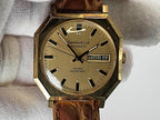 A vintage Caravelle Day-Date wristwatch with an octagonal gold-tone case, beige dial, and brown leather strap, held against a white textured background.