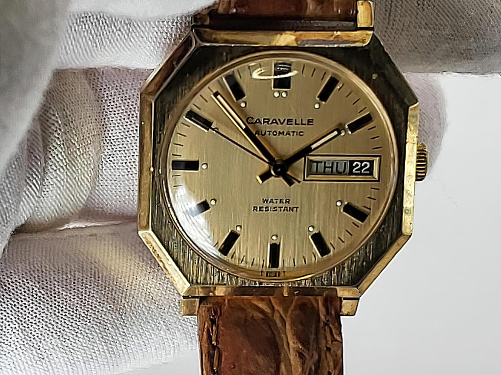 A vintage Caravelle automatic watch with an octagonal gold-tone case, cream dial, and brown leather strap, held against a white textured fabric background.