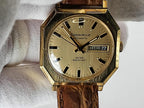 A vintage Caravelle automatic watch with an octagonal gold-tone case, cream dial, and brown leather strap, held against a white textured fabric background.