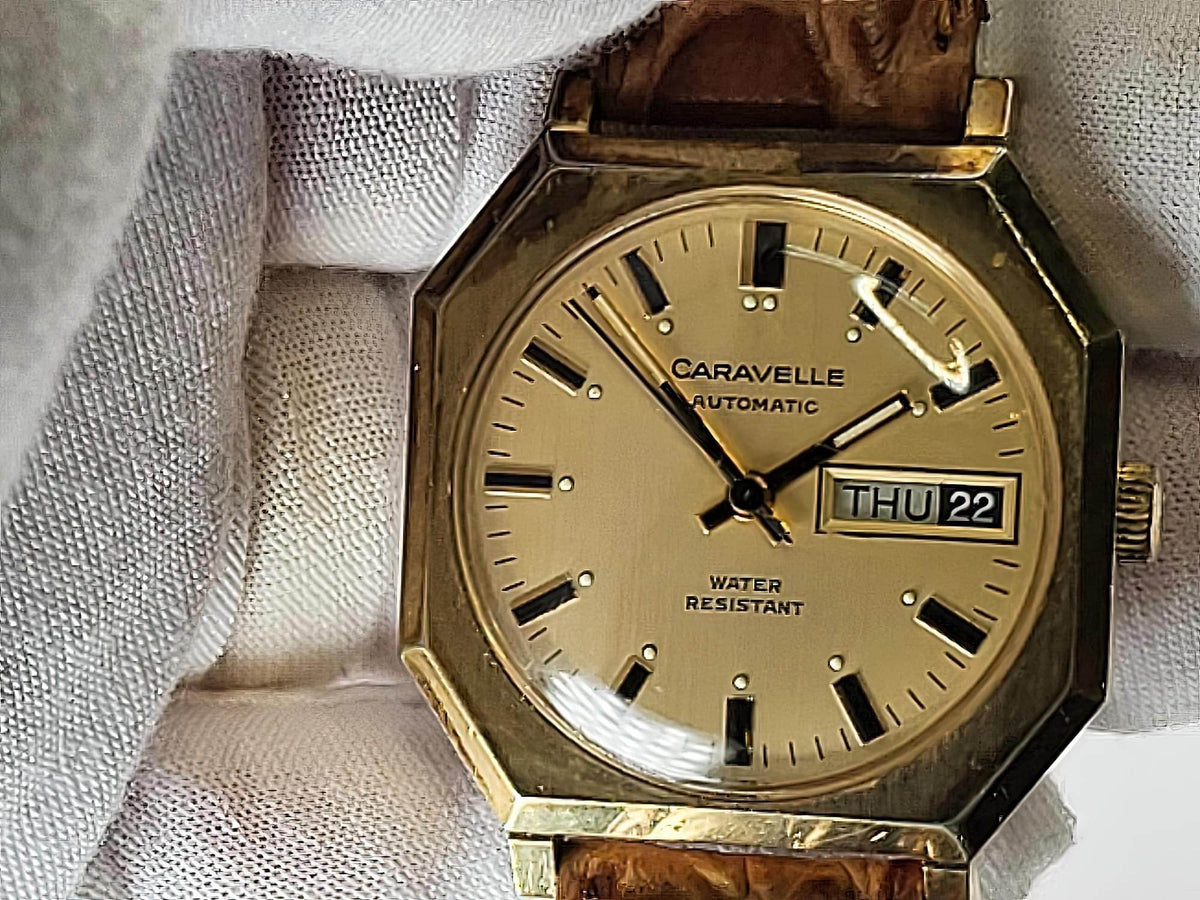 A vintage Caravelle automatic wristwatch with an octagonal gold-tone case, beige dial, and brown leather strap, resting on a white textured fabric surface.