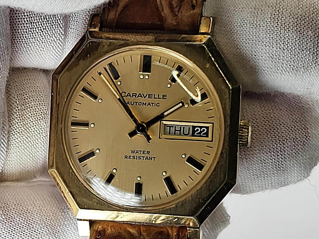 A vintage Caravelle automatic wristwatch with an octagonal gold-toned case, beige dial, black hands and markers, and a brown leather strap, resting on a white textured fabric.