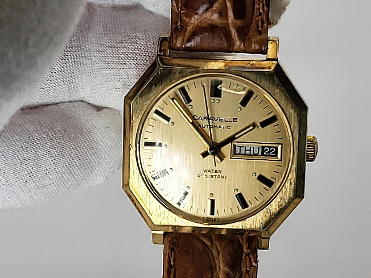 A vintage Caravelle automatic wristwatch with an octagonal gold-tone case, cream dial, and brown leather strap, held by a white-gloved hand against a light gray background.