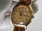 A vintage Caravelle automatic wristwatch with an octagonal gold-tone case, beige dial, and brown leather strap, held by a white gloved hand.