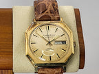 A vintage Caravelle Day-Date automatic wristwatch with an octagonal gold-tone case, cream dial, and brown leather strap, displayed on a beige cushion mounted on a wooden block.