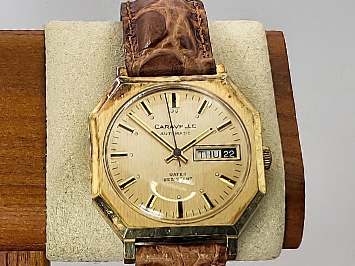 A vintage Caravelle Day-Date automatic wristwatch with an octagonal gold-tone case, cream dial, and brown leather strap, displayed on a beige cushion mounted on a wooden block.