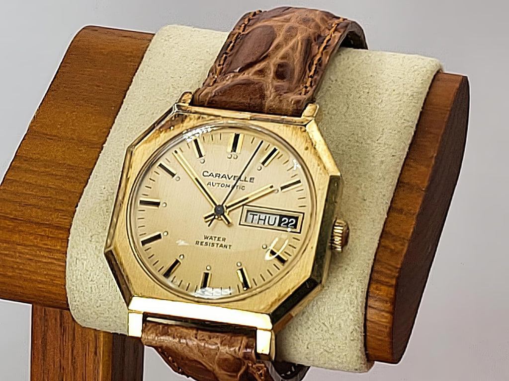 A vintage Caravelle Day-Date wrist watch with a gold-tone octagonal case, beige dial, and brown leather strap, displayed on a wooden stand with a beige cushion.