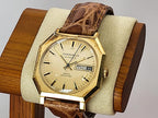 A vintage Caravelle Day-Date wrist watch with a gold-tone octagonal case, beige dial, and brown leather strap, displayed on a wooden stand with a beige cushion.