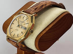 A vintage Caravelle Day-Date wristwatch with a gold-tone case, cream dial, and brown leather strap, displayed on a wooden stand with a cream cushion against a light gray background.