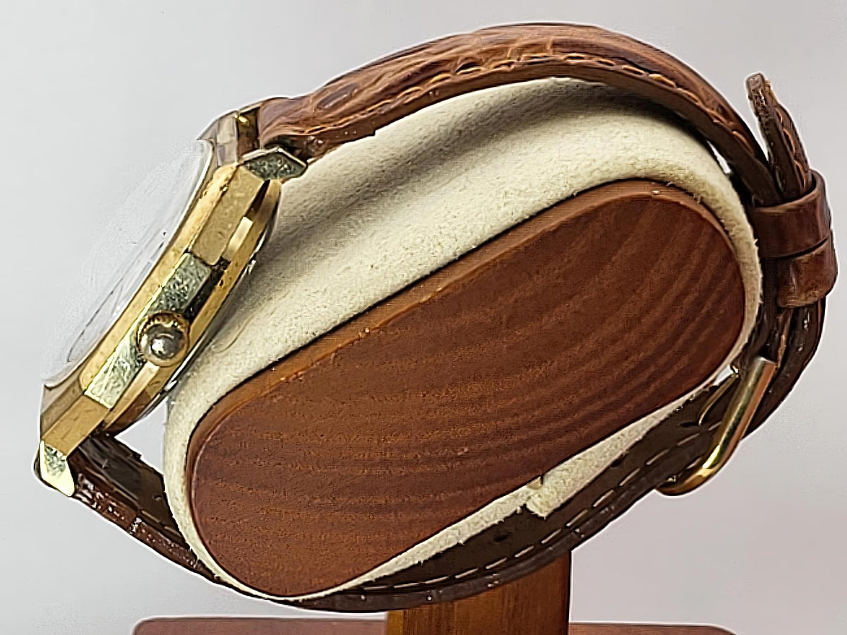 A close-up of a vintage Caravelle Day-Date watch with a brown leather strap and gold-toned case, displayed on a wooden stand against a light gray background.