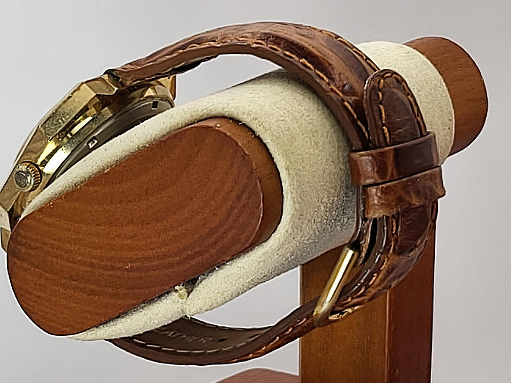 A close-up of a brown leather Caravelle Day-Date watch strap with gold-toned hardware, draped over a cylindrical display stand with a cream-colored velvet interior.