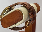A close-up of a brown leather Caravelle Day-Date watch strap with gold-toned hardware, draped over a cylindrical display stand with a cream-colored velvet interior.