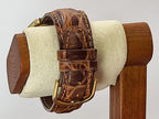 A brown leather watch strap for a Caravelle Day-Date wristwatc, with a textured alligator pattern and gold-tone buckle, displayed on a cream-colored velvet stand with a wooden base.