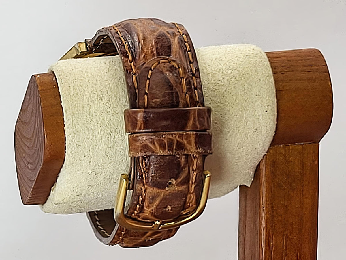 A brown leather watch strap for a Caravelle Day-Date wristwatc, with a textured alligator pattern and gold-tone buckle, displayed on a cream-colored velvet stand with a wooden base.