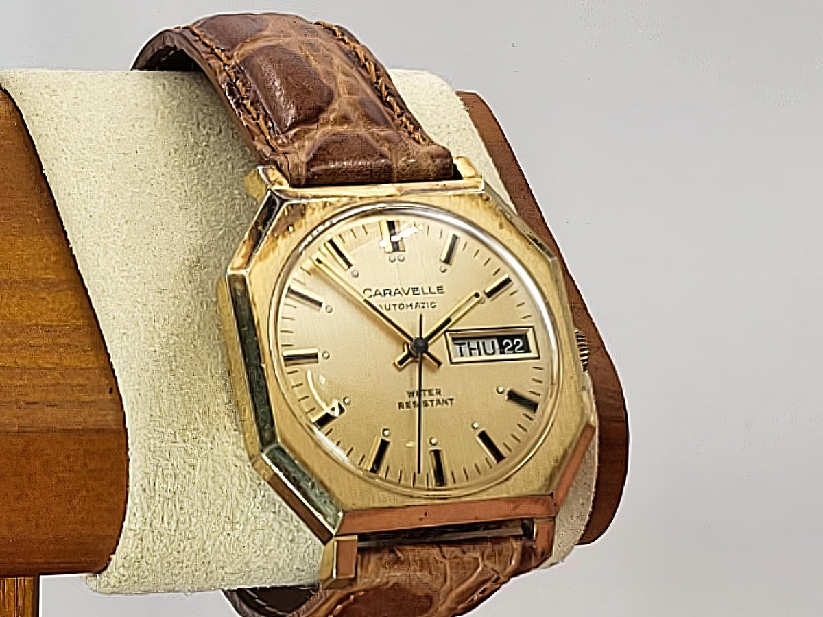 A vintage Caravelle Day-Date  wristwatch with an octagonal gold-tone case, cream dial, and brown leather strap, displayed on a cream cushion mounted on a wooden stand.